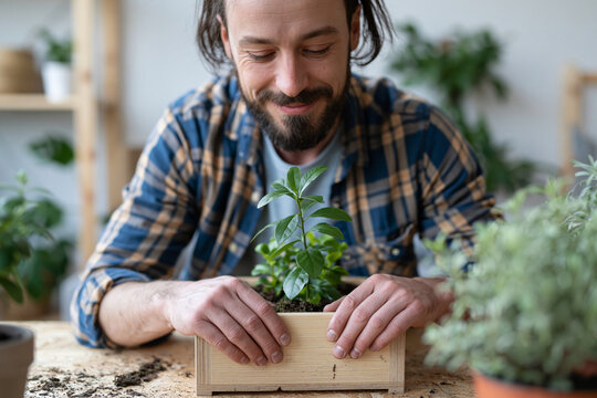 Man smiles gently while tending a small green plant. Fingers press soil around young leaves in a wooden box. Background holds soft-focus greenery and shelves. Natural light highlights warmth
