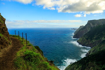 Fototapeta premium Machico, Portugal - 03.12.2025: Narrow dirt hiking trail with rope fence overlooking steep green cliffs and deep blue Atlantic Ocean