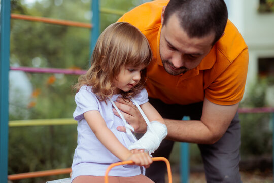 Caring Father Helping Little Daughter With Bandaged Arm At Playground. A worried parent supports his injured girl with a broken hand outdoors 