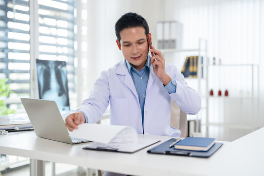 Doctor in telemedicine concept. asian doctor using phone while writing notes and working on  laptop at the medical office.
