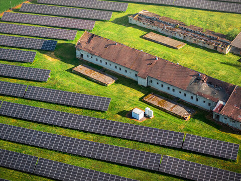 Aerial view of a large solar farm with rows of photovoltaic panels installed on green grass next to an old, weathered farm building in Chvalkovice, Hradec Kralove Region, Czechia.