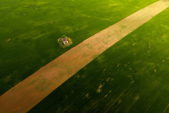 Aerial view of a small, weathered structure standing alone in a vast green field, bisected by a diagonal strip of brown earth under soft light in Cejkovice, South Moravian Region, Czechia.
