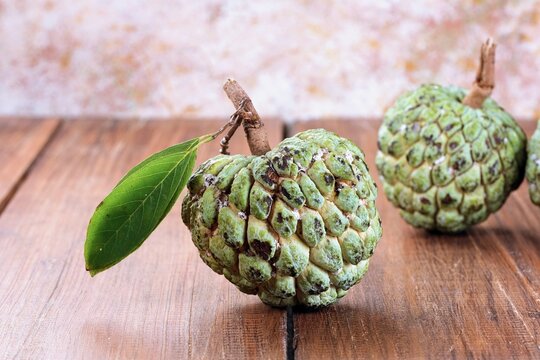 Ripe atemoya custard apple fruits displayed with green leaves on a rustic surface, showing their distinctive segmented texture and natural tropical appearance.