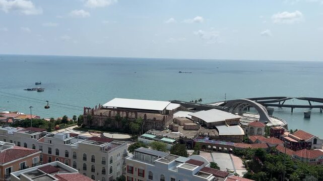 4k footage of panoramic view of a coastal town by the blue sea. Colorful buildings and a large arena sit near the water, with a cable car and boats moving along the clear ocean under a sunny sky