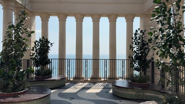 4k footage walking through classic roman stone pillars on a balcony. Green potted plants and whole pillars opening up to a wide view of the blue ocean under a bright sky.