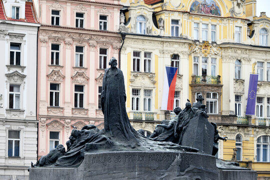 Bronze figures of the Jan Hus Memorial with a pink building and Czech flag in the background, Old Town Square, Prague, Czech Republic