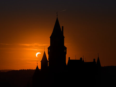 Drone photo of Bouzov Castle in silhouette during a partial solar eclipse at sunset, with the dark castle towers outlined against a glowing orange evening sky in the Czech Republic. 