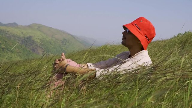 Man in orange hat sitting in grassy meadow in India, enjoying nature and mountain views.
