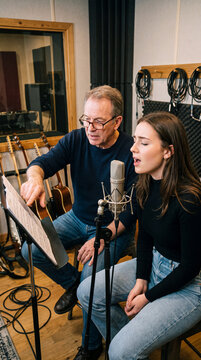 Vocal coach guides young singer in recording studio with microphone and music sheet