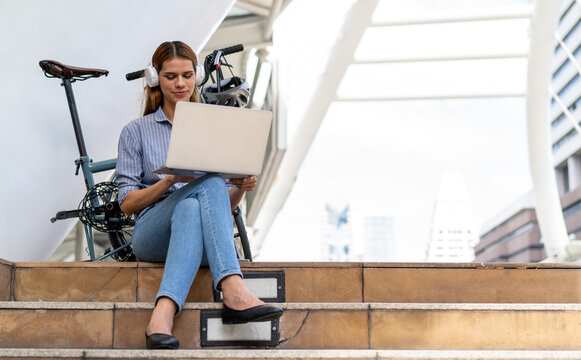 Urban Nomad with Laptop and Bicycle: A young woman with headphones sits on steps outside a modern building, working on her laptop with a folded bicycle beside her. Bike to work.