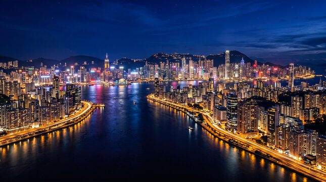 Illuminated skyscrapers stretch across dark coastal horizon displaying vibrant neon lights reflecting upon calm harbor waters during peaceful evening aerial cityscape view