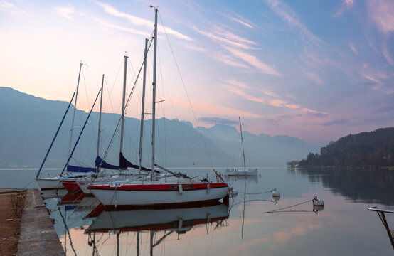 Sailboats reflected in the calm water of Lake Annecy at sunrise with mountains in the background, France.
