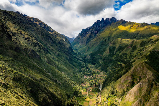 Aerial view of the Sacred Valley in the Andes mountains with lush green slopes, agricultural fields, and a small settlement nestled in the valley floor, Urubamba Province, Cusco, Peru.