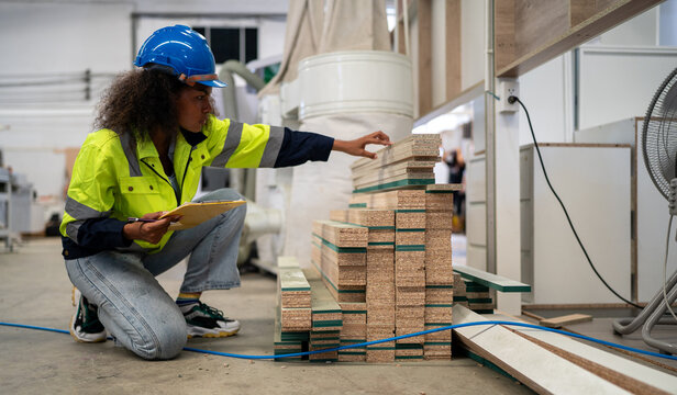Warehouse Inspection: A diligent black female worker in a hard hat and safety vest crouches by a stack of lumber, meticulously recording details on a clipboard. Multiracial woman worker.