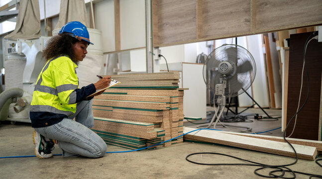 Warehouse Inspection: A diligent black female worker in a hard hat and safety vest crouches by a stack of lumber, meticulously recording details on a clipboard. Multiracial woman worker.