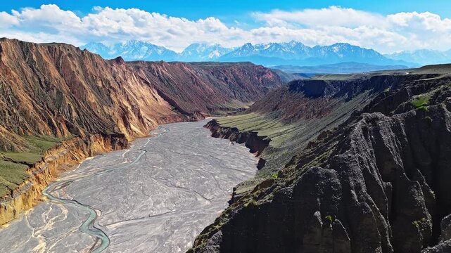 Aerial view of a deep canyon with a braided riverbed and snow-capped mountains under a blue sky in Anjihai Grand Canyon, Xinjiang, China