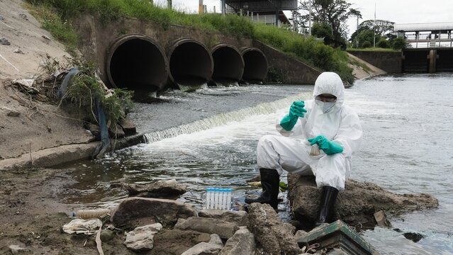 Environmental Specialist in Hazmat Suit Testing Water Quality Near Concrete Drain Pipes Wide Shot