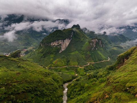 Aerial view of the Ha Giang Loop winding through lush green karst mountains and valleys with a river flowing below under a cloudy sky in Dong Van, Tuyen Quang, Vietnam.