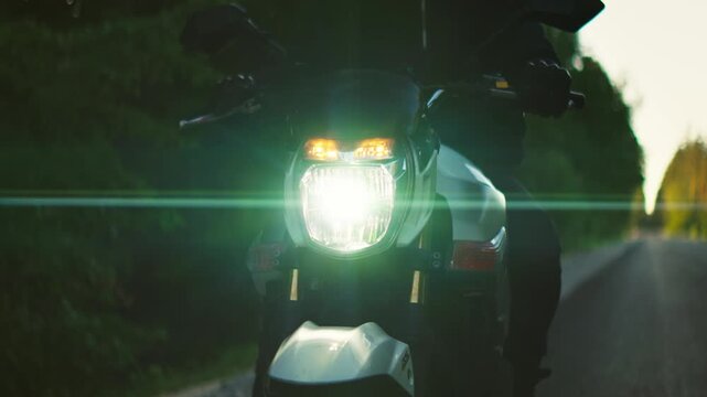 Motorcycle headlight turning on at dusk on a forest road, illuminating the dark surroundings as a lone rider prepares for a night journey along a quiet highway