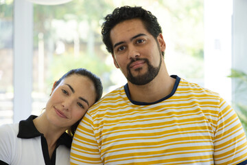 Fototapeta premium Diverse couple standing close in bright room by windows, wearing striped T-shirt and collared top