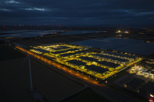 Aerial view of a large hyperscale AI data center illuminated at night, featuring rows of industrial buildings, surrounding dark agricultural fields, and a wind turbine.