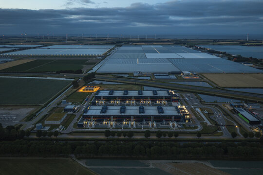 Aerial view of a hyperscale AI data center illuminated at dusk, surrounded by expansive glass greenhouses and distant wind turbines under a twilight sky.