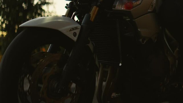 Sport motorcycle riding along a quiet rural road at golden hour, closeup of spinning front wheel and illuminated headlight against warm sunset landscape