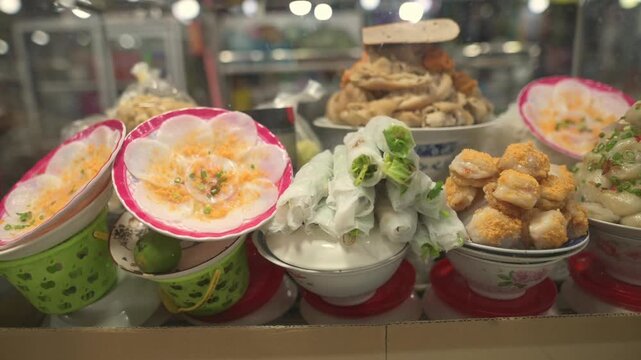 Vietnamese Street Market Dessert Display With Colorful Bowls Of Coconut Pudding, Dragonfruit Halves, Fried Tofu Cubes And Steamed Rice Rolls. Vendor Arranging Bowls Behind Glass Case, Warm