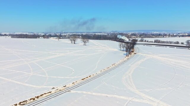 A steam passenger train moves slowly through a snowy field. The bright sky adds to the scene. Snow covers the ground and trees. Smoke comes from the train as it progresses along the track.