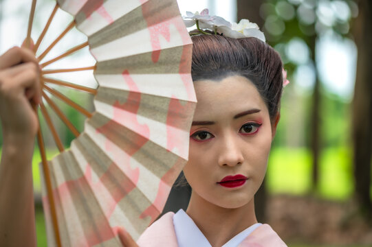 Portrait young Japanese geisha in a delicate pastel kimono holding a traditional fan covering face enjoying vibrant green garden 
