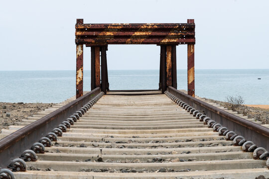 Old railway tracks ending at rusty buffer stop with calm blue ocean view in Sri Lanka. Symmetry shot of train rails leading towards the sea coastline under clear sky.