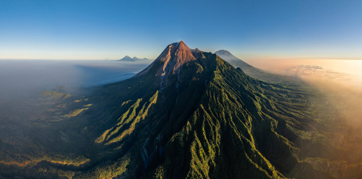 Aerial view of the majestic Mount Merapi volcano with lush green slopes and rugged volcanic terrain under a clear blue sky in Cangkringan, Special Region of Yogyakarta, Indonesia.