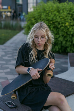 Blonde teenager inspects their skateboard on a bench