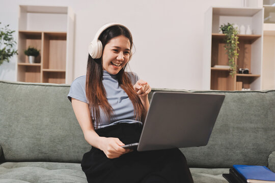 Beautiful young asian woman sitting on sofa and holding joystick playing online video game in living room at house