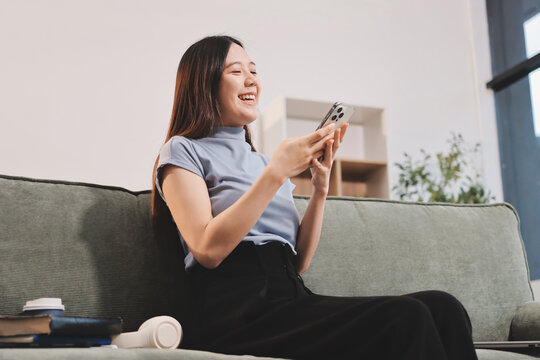 Freelance Asian woman holding mobile phone typing on laptop and working from home. Happy young girl sitting on sofa in living room.