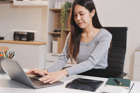 Young asian businesswoman is smiling and working on a laptop while sitting at her desk in a modern office