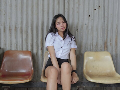 A young Asian woman with long straight black hair, wearing a white short-sleeved collared shirt and black skirt, sits on a worn beige plastic chair against a rusty corrugated metal wall