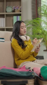 Asian woman writing checklist before packing suitcase for vacation trip