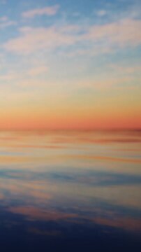 The sun sets over a large body of water. The sky shows shades of orange and purple. The water is smooth and mirrors the colors from above. It is evening time and the scene is quiet.