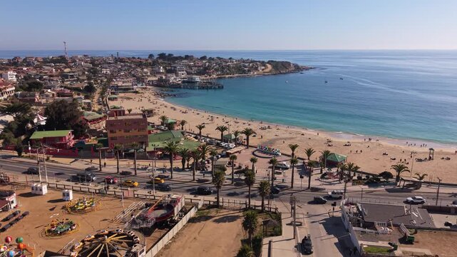 Advancing flight over beachfront boardwalk with amusement rides and palm lined avenue revealing the crescent shaped bay and turquoise ocean waters. El Quisco, Chile