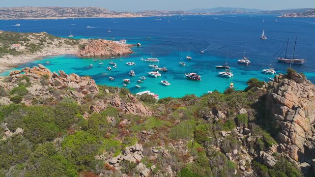 Epic aerial flying over granite cliffs to reveal luxury sailing yachts in crystal clear water of a natural protected harbor on the Isola di Spargi in the Maddalena Archipelago in Sardinia, Italy.