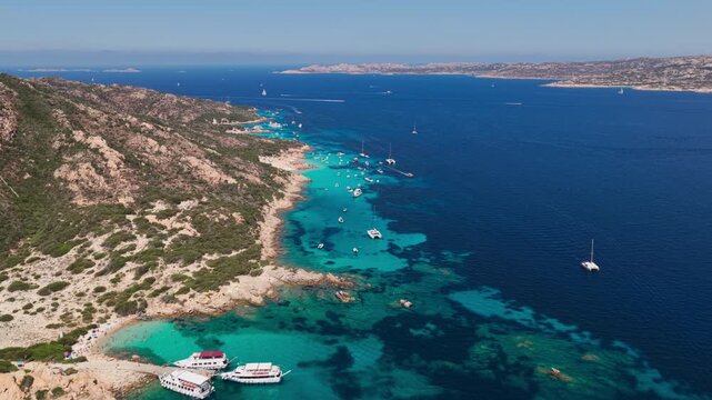 Epic aerial flying over luxury sailing yachts in a strait of turquoise blue water, past granite cliffs and the rocky coastline of the Isola di Spargi in the Maddalena Archipelago in Sardinia, Italy.