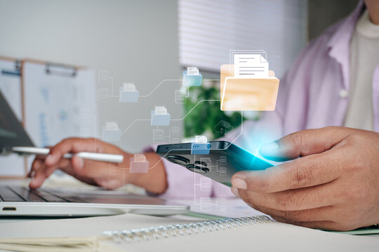 Person using smartphone and laptop at desk with digital document management system holographic graphic, concept of cloud storage, electronic filing, data organization, and paperless office.