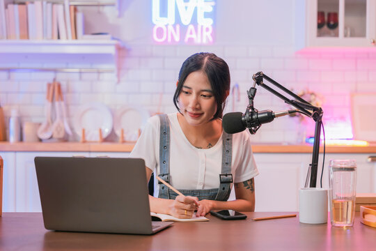Focused young Asian woman concentrating on her laptop while working from a home podcast studio, with a glowing "Live On Air" neon sign and microphone in the background.