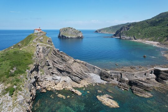Aerial view of the hermitage of Gaztelugatxe on a rocky islet connected by a stone bridge to the mainland, surrounded by the blue waters of the Bay of Biscay in Basque Country, Spain.