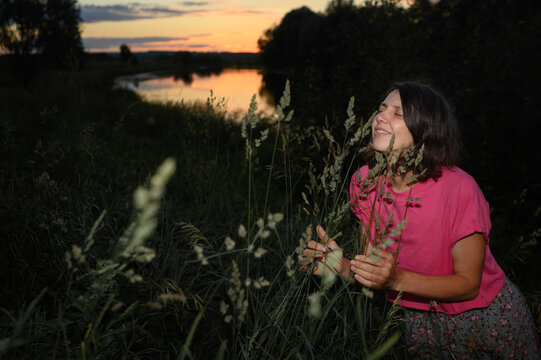 Woman smells tall grass near a river during a peaceful summer sunset