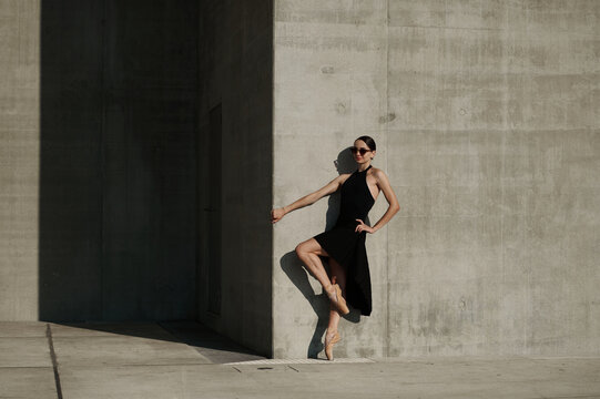 Elegant female ballet dancer in black dress and sunglasses posing on pointe against minimalist concrete architecture in sunlight.