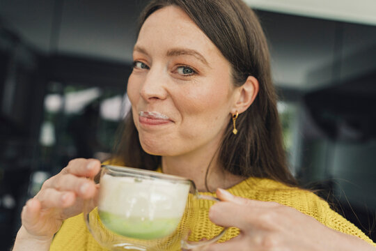 Woman drinks homemade matcha latte with milk beard at home