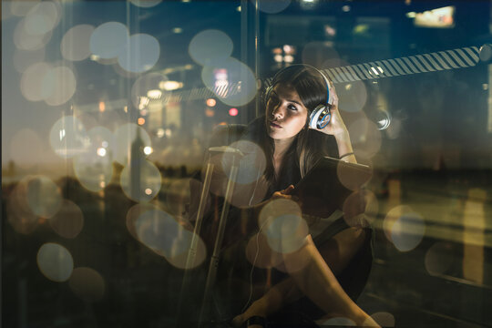 Young woman with tablet and headphones sitting outdoors at night