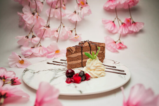 Delicious chocolate cake slice served with whipped cream and fresh berries on a white marble plate surrounded by sakura